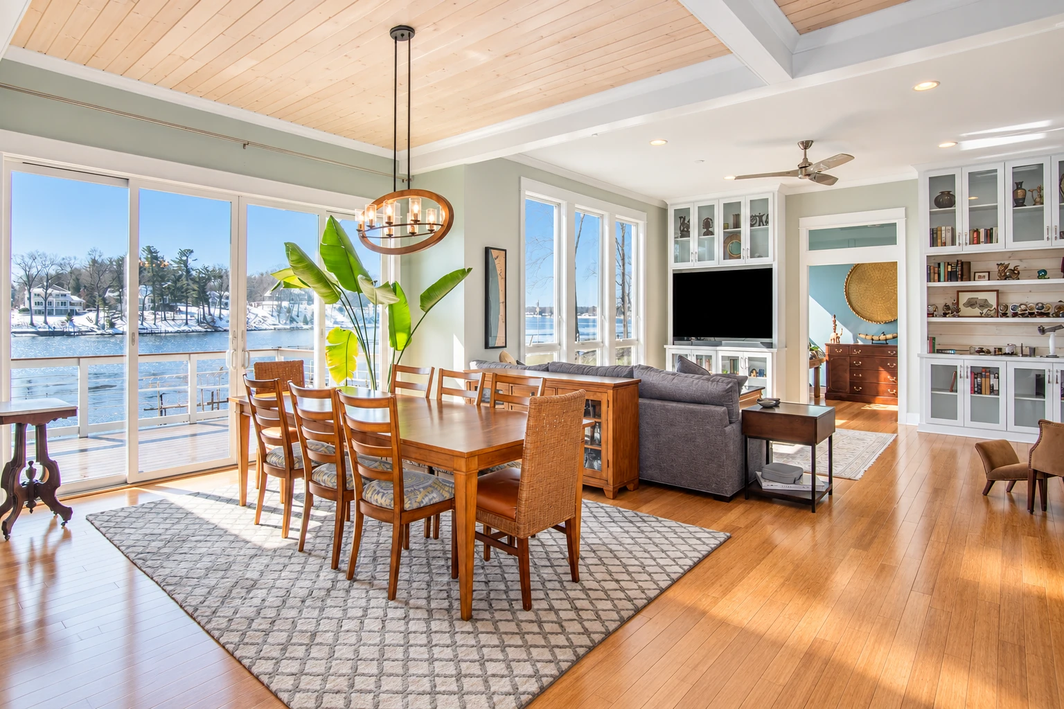 Sunroom with large windows overlooking a lake, featuring a wood dining table, vaulted wood ceiling, and adjoining living area.
