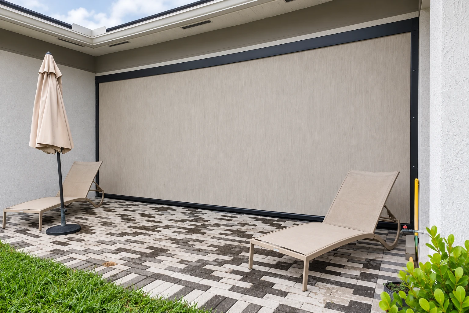 A covered patio has a large lowered beige screen, two chaise lounges, a closed umbrella, and a patterned paver floor.