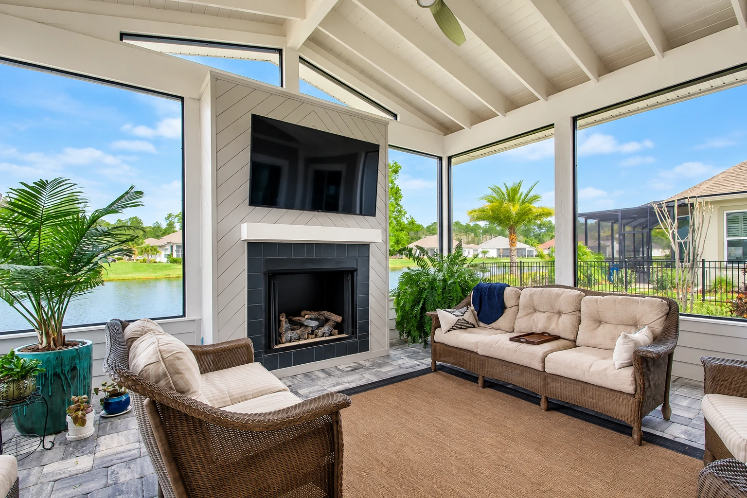 Sunroom addition with wicker seating, a fireplace and TV, surrounded by large windows overlooking a lake.