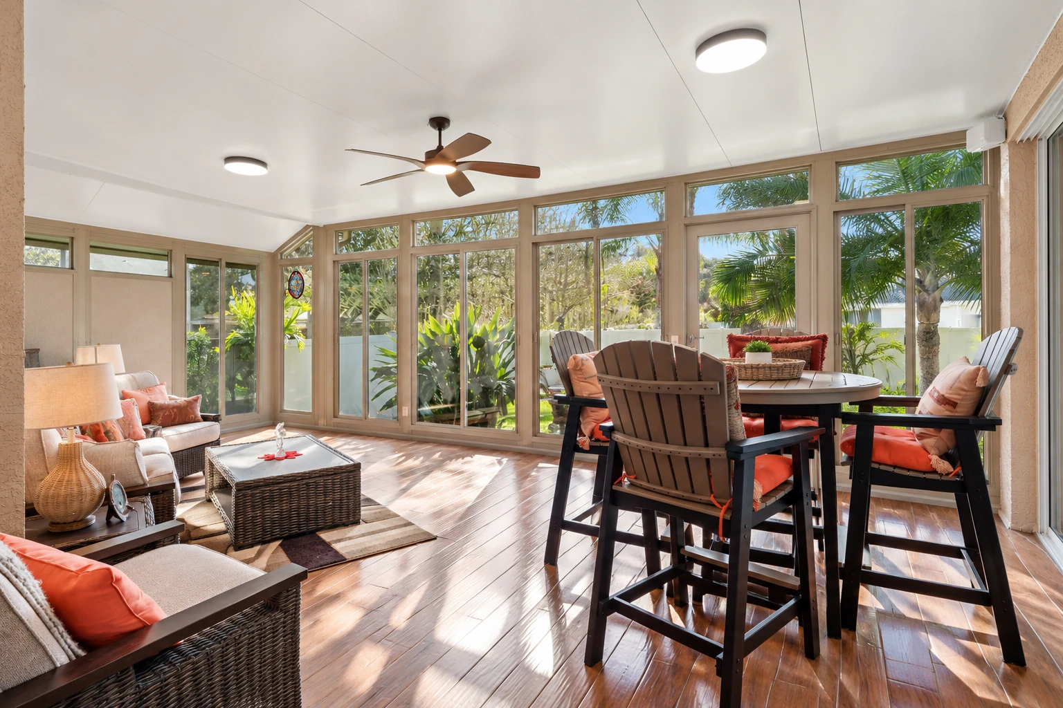 Bright sunroom with large windows, wood-look flooring, lounge seating, and a dining table surrounded by greenery.