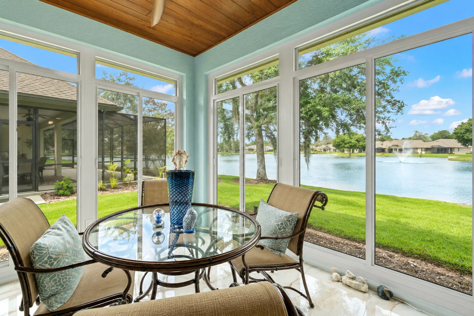 Sunroom with a wood-paneled ceiling and broad windows overlooking a lake, furnished with a glass dining table and wicker chairs.