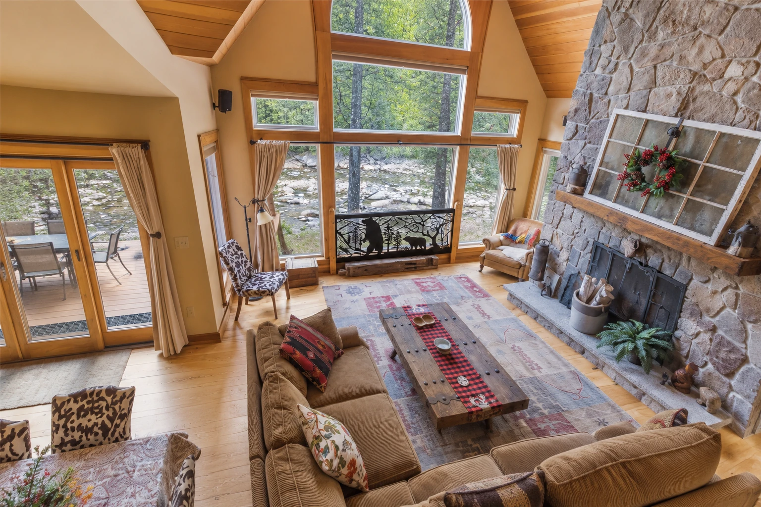 Sunroom with tall wood-framed windows, vaulted ceiling, stone fireplace, and rustic seating.