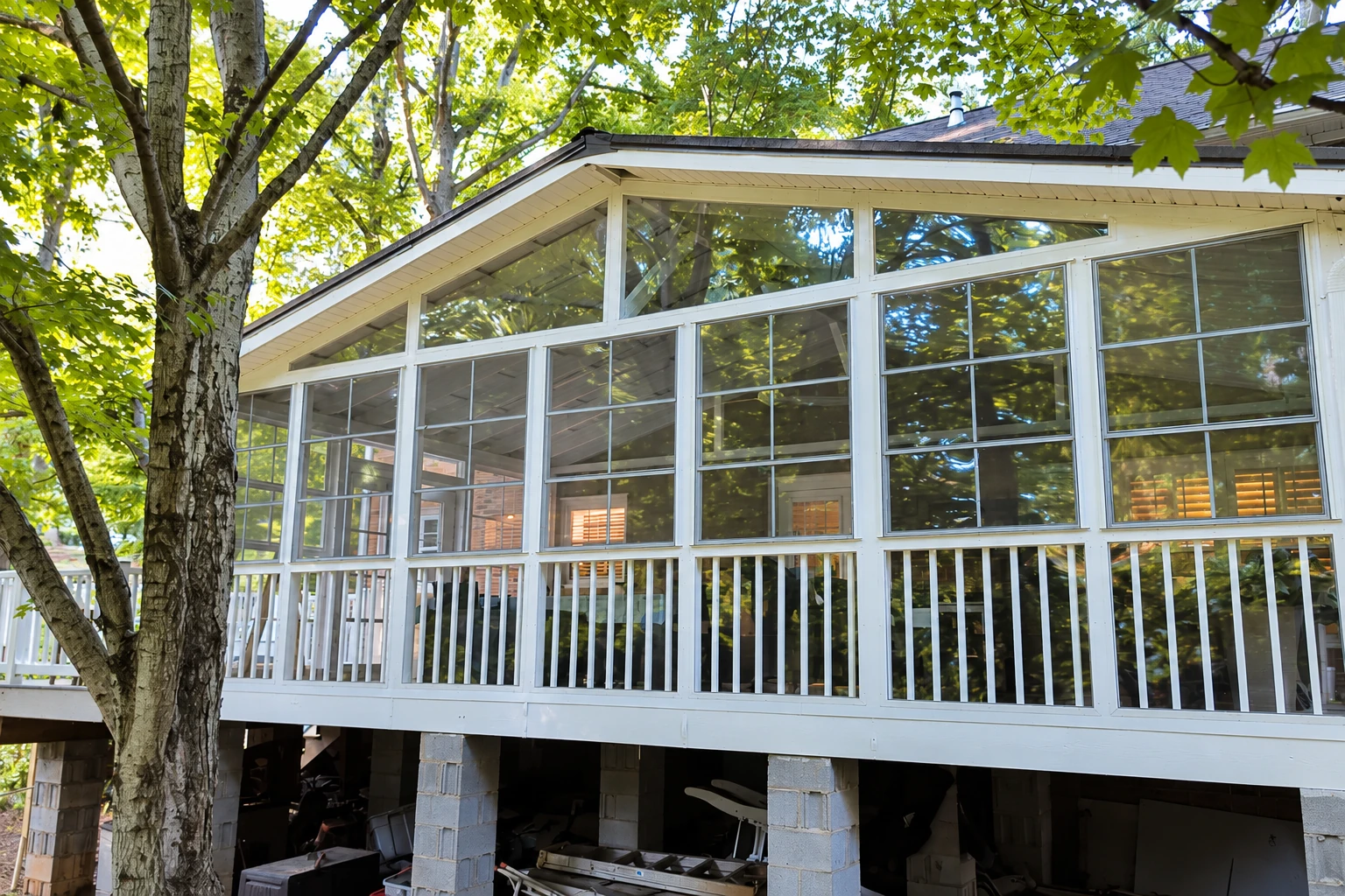 Three-season sunroom with white-framed windows and screened lower panels beneath leafy trees.