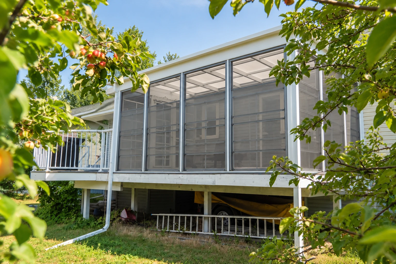 Raised three-season sunroom with screened walls and a white frame, partly surrounded by leafy branches.